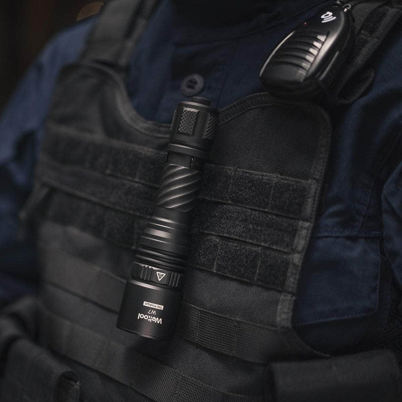 A person wearing a tactical vest with a Weltool W7 long-range LEP tactical flashlight (700 lumens) clipped to the front and a radio attached to the shoulder.