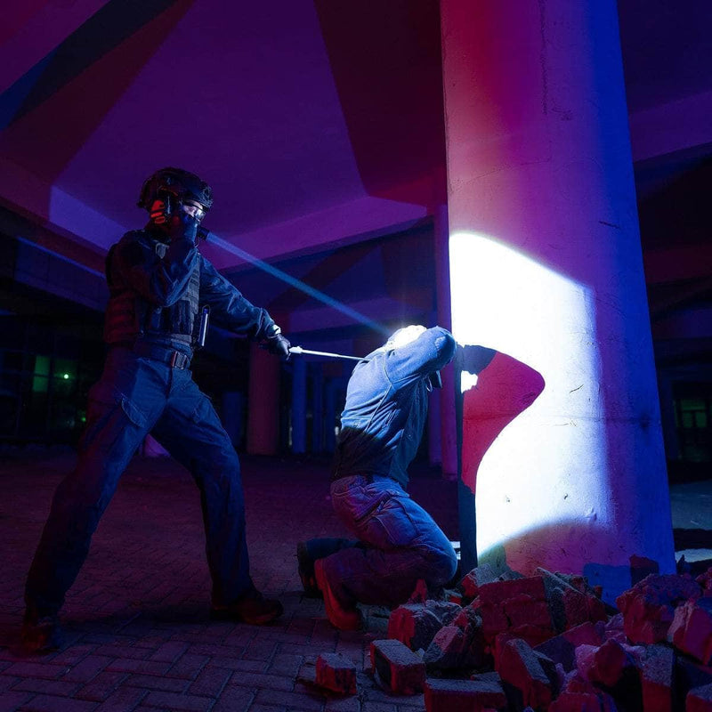 A police officer holds a baton and shines a Weltool W7 long-range LEP tactical flashlight on a kneeling person by a pillar in a dimly lit area with scattered bricks.