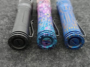 Three distinct ReyLight Anodized Ti LAN flashlights—a plain black, a pink-blue camouflage, and a blue circuit pattern—are showcased side by side on a gray surface, embodying sleek elegance and preparing to illuminate any adventure.