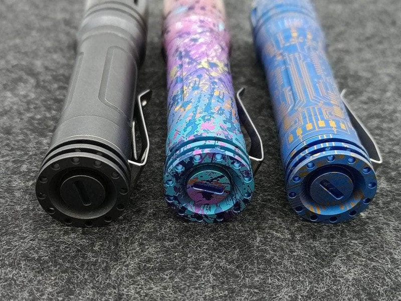 Three distinct ReyLight Anodized Ti LAN flashlights—a plain black, a pink-blue camouflage, and a blue circuit pattern—are showcased side by side on a gray surface, embodying sleek elegance and preparing to illuminate any adventure.