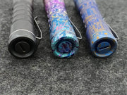 Three uniquely designed pens and a sleek ReyLight Anodized Ti Pineapple flashlight are placed horizontally on a textured gray surface.