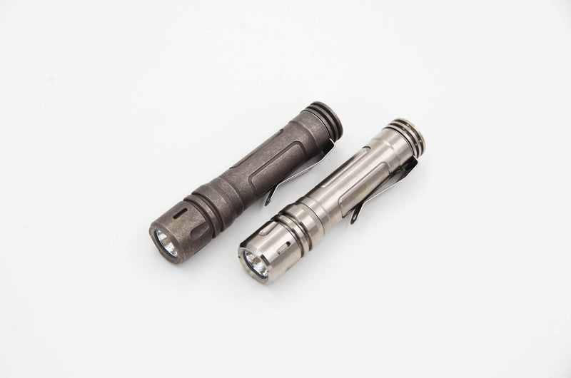Two tactical flashlights, one dark and one metallic, with pocket clips rest on a white background. The sleek design of the ReyLight LAN Titanium with Nichia 219B LED offers an efficient and reliable companion for any adventure.