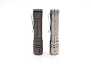 Two metallic flashlights with pocket clips stand upright on a white background, one in dark gray and the other in silver. The ReyLight LAN Titanium model, featuring a titanium design and Nichia 219B LED, offers style and superior luminosity.