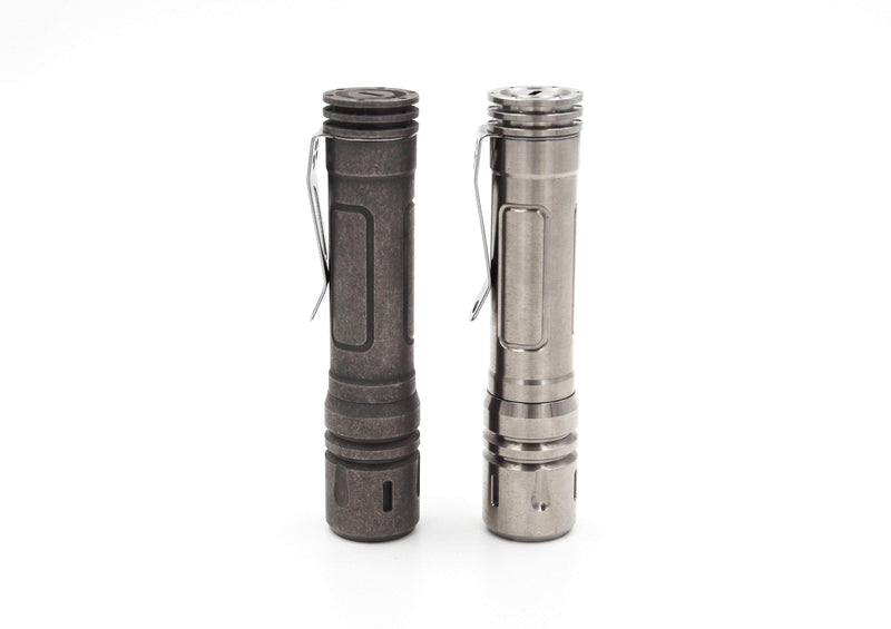 Two metallic flashlights with pocket clips stand upright on a white background, one in dark gray and the other in silver. The ReyLight LAN Titanium model, featuring a titanium design and Nichia 219B LED, offers style and superior luminosity.