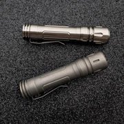 Two metallic flashlights with clips lie parallel on a textured black surface, including a ReyLight LAN Titanium featuring the Nichia 219B LED.