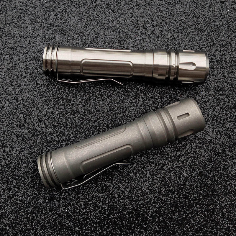 Two metallic flashlights with clips lie parallel on a textured black surface, including a ReyLight LAN Titanium featuring the Nichia 219B LED.