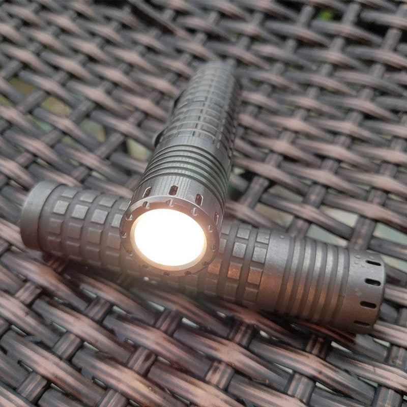 Two ReyLight Ti Dawn Triple tactical flashlights, featuring Nichia 519A emitters, rest on a woven surface. Their glow highlights exquisite craftsmanship with sapphire lenses and sleek designs.