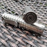 A ReyLight Ti Dawn Triple, a silver metal flashlight with a textured grip and pocket clip, sits on a woven surface. Its end cap has a circular button, while its Sapphire lens and Nichia 519A emitters illuminate the path ahead.